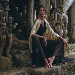 May include: A woman wearing a beige silk kimono and black trousers sits on a stone step in front of two wooden carvings. The carvings are of human figures with elaborate headdresses.