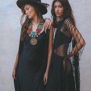 May include: Two women wearing black dresses. One woman is wearing a wide-brimmed black hat and a colorful beaded necklace with a large metal pendant. The other woman is wearing a black mesh dress with fringe.