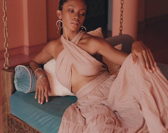 May include: A woman wearing a light pink halter top and matching skirt sits on a wooden swing with a teal cushion. She has dark hair and is wearing silver hoop earrings.