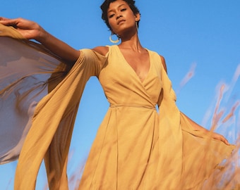 May include: A woman wearing a long, flowing, mustard yellow dress with a wrap style bodice and long sleeves. The dress is made of a lightweight fabric and has a flowing, ethereal look. The woman is standing in a field of tall grass, and the wind is blowing her hair and dress.