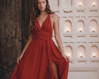 May include: A woman wearing a long, rust-colored maxi dress with a plunging neckline and a braided rope belt. The dress has a thigh-high slit and is made of a lightweight fabric.