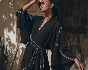 May include: A black kimono with gold embroidery and a long, flowing silhouette. The kimono is worn over a red skirt and the model is wearing a black hat.