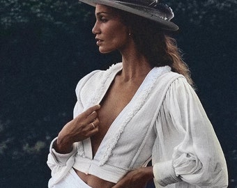 May include: A woman wearing a white, long-sleeved,  cropped top with a deep V-neckline and a white wide-leg pant. She is wearing a white fedora with a black feather.