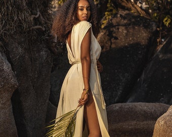 May include: A woman wearing a flowing, pale green dress with a high slit, holding a palm leaf. The dress has a low back and is cinched at the waist. She is standing on a sandy beach with large rocks in the background.