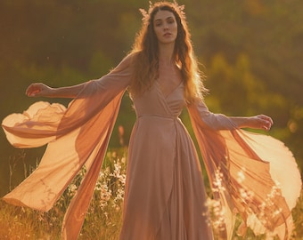 May include: A woman wearing a long, flowing, light brown dress with long sleeves and a flowing skirt. She is standing in a field of tall grass with the sun shining on her.