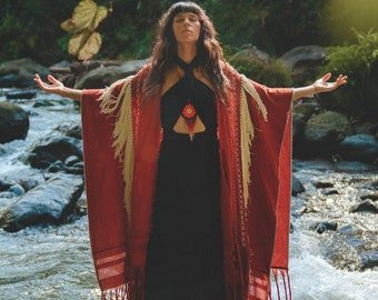 May include: A woman wearing a black dress and a red shawl with cream-colored fringe stands in a river. The shawl has a geometric pattern and is draped over the shoulders. The woman has long dark hair and is standing with arms outstretched.
