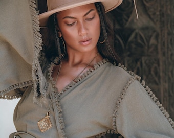 May include: A woman wearing a sage green wrap top with fringe detailing and a brown fedora. She is wearing a gold necklace with a pendant. The top has a tie at the waist.