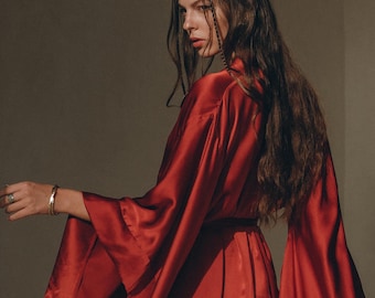 May include: A deep red satin robe featuring long, wide bell sleeves and a wrap-around design with a tie waist. The garment has a smooth, reflective finish and a fluid drape, showcasing a vibrant crimson hue throughout the entire piece.