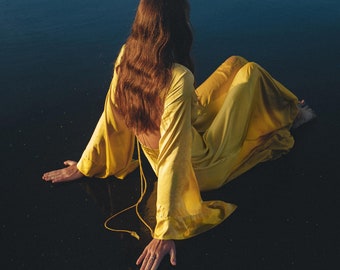 May include: A woman wearing a yellow dress sits on a black sand beach, facing the sea. The waves are breaking in the background.