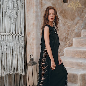 May include: A woman wearing a black maxi dress with fringe detailing. The dress has a sleeveless design and a relaxed fit. The woman is standing on a wooden deck with a stone wall and a macrame wall hanging in the background.