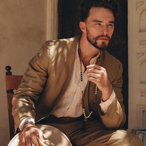 以下が含まれることがあります： 薄い茶色のスーツジャケットとパンツを着た男性が椅子に座っています。彼は白いボタンダウンシャツとビーズネックレスをしています。彼は薄い茶色のフェドーラハットを膝の上に置いています。