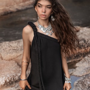 May include: A woman with long brown hair wearing a black one-shoulder dress with a rope detail. She is wearing silver jewelry, including a necklace, earrings, and bracelets.