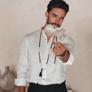 以下が含まれることがあります： 白いリネンシャツにビーズネックレス、黒いパンツを着用した男性が、白い花を前に持っている。