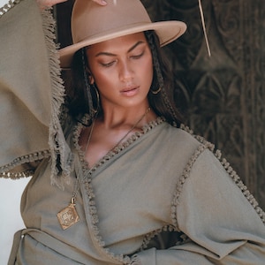 Puede incluir: Una mujer con un top verde salvia de envoltura con detalles de flecos y un fedora marrón. Lleva un collar de oro con un colgante. El top tiene un lazo en la cintura.