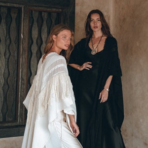 May include: Two women wearing long, flowing garments. The woman on the left is wearing a white, fringed poncho. The woman on the right is wearing a black, long-sleeved dress with a silver pendant necklace.