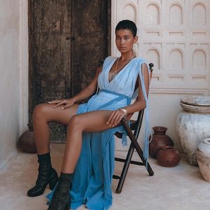 May include: A woman wearing a light blue maxi dress with a silver necklace and bracelets sits on a wooden chair in front of a white wall with decorative arches. She is wearing black heeled boots.