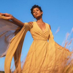 Puede incluir: Una mujer lleva un vestido largo, fluido, de color amarillo mostaza con un corpiño de estilo envolvente y mangas largas. El vestido está hecho de una tela ligera y tiene un aspecto fluido y etéreo. La mujer está de pie en un campo de hierba alta, y el viento le está soplando el pelo y el vestido.
