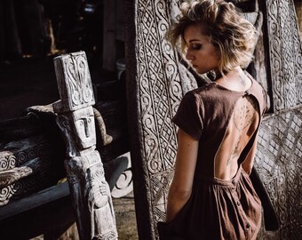 May include: A woman with short blonde hair wears a brown dress with an open back. She stands in front of a carved wooden wall with intricate designs. The wall is made of several panels with different patterns.