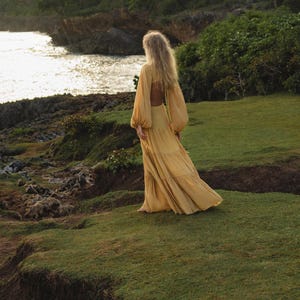 A woman in the Samudra Dress in Saffron Gold by AYA Sacred Wear, a cotton maxi with a tiered bohemian silhouette and waist cut-outs, stands on a grassy cliff at sunset, surrounded by lush greenery and rocky terrain as she overlooks the ocean.