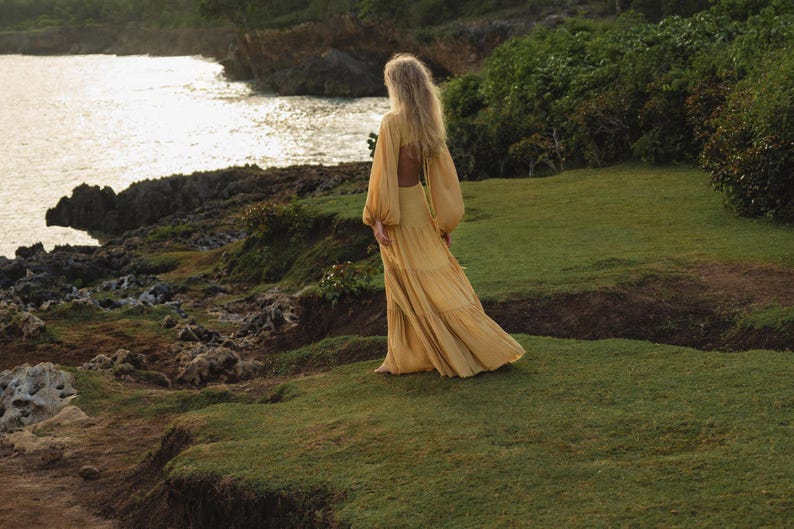 A woman in the Samudra Dress in Saffron Gold by AYA Sacred Wear, a cotton maxi with a tiered bohemian silhouette and waist cut-outs, stands on a grassy cliff at sunset, surrounded by lush greenery and rocky terrain as she overlooks the ocean.