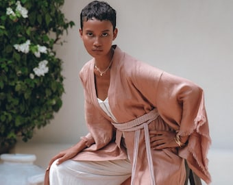 May include: A woman wearing a light peach-colored kimono-style robe with fringed sleeves, a woven belt, and white pants. The robe is open, revealing a white top. The model is wearing gold bracelets and a delicate necklace. The background is a white wall with greenery.