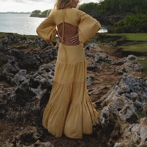 A woman in the AYA Sacred Wear Samudra Dress in Saffron Gold—a cotton maxi with a tiered bohemian silhouette and waist cut-outs—stands on rocky coastal terrain, gazing at the sea amid lush greenery and a cloudy sky.