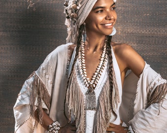 May include: A woman wearing a cream-colored outfit with shell and beaded jewelry. The ensemble includes a head wrap, necklace, earrings, and bracelets. The dress has fringe and embroidered details. The overall style is bohemian.