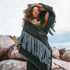 May include: A woman with curly hair wears a black shawl with fringe and a white shell necklace. The shawl has a geometric pattern of white stitching. The woman is standing in front of large rocks.