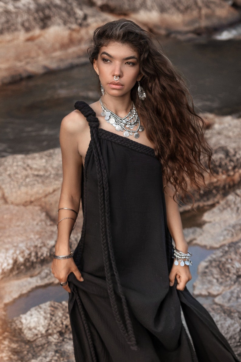 May include: A woman with long brown hair wearing a black one-shoulder dress with a rope detail. She is wearing silver jewelry, including a necklace, earrings, and bracelets.