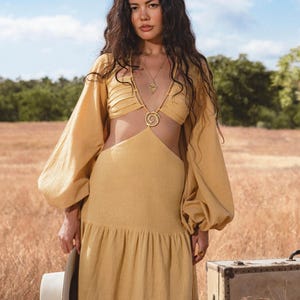 A woman with long wavy hair stands in a dry grassy field, wearing the AYA Sacred Wear Samudra Dress in Saffron Gold—a cotton maxi dress featuring a tiered bohemian silhouette and waist cut-outs. She holds a white hat by a vintage suitcase under the blue sky.