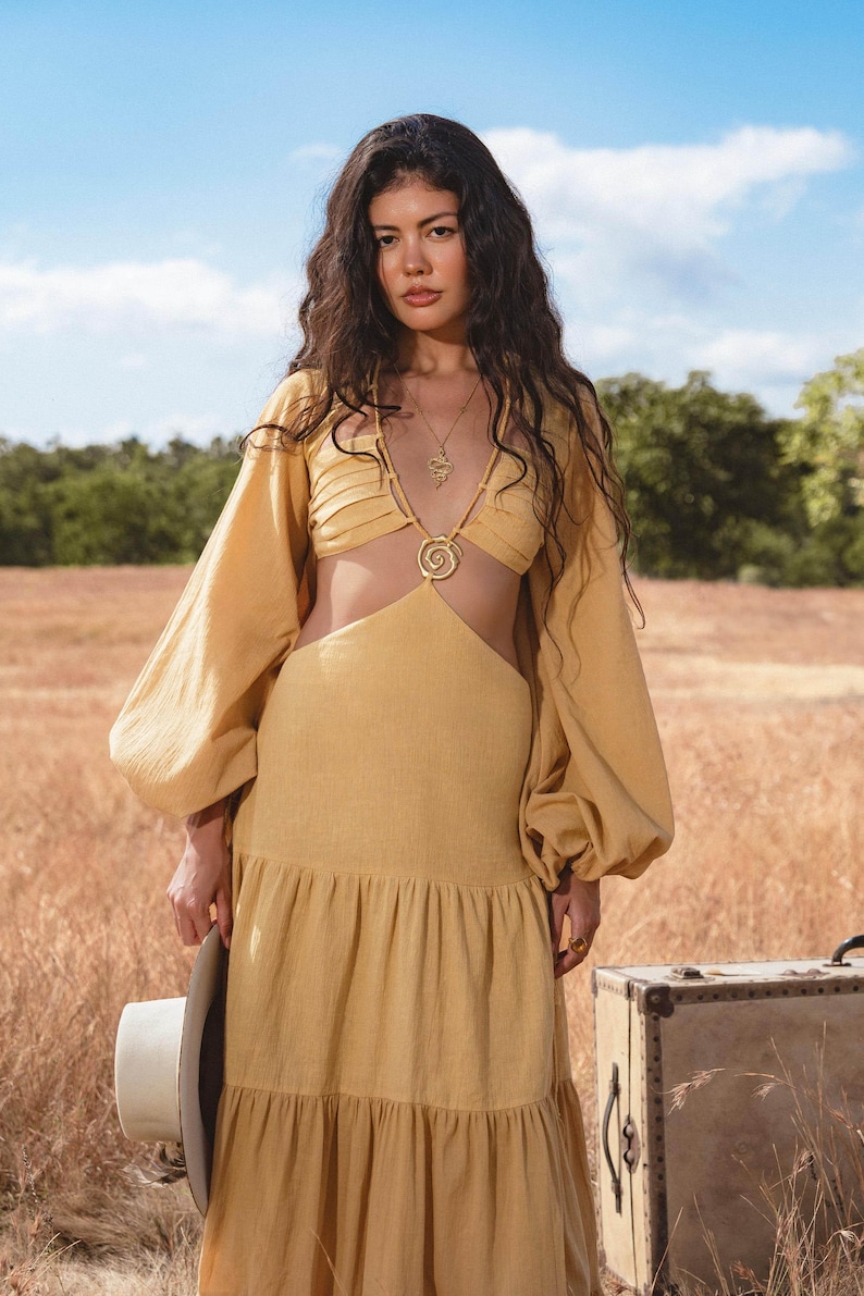 A woman with long wavy hair stands in a dry grassy field, wearing the AYA Sacred Wear Samudra Dress in Saffron Gold—a cotton maxi dress featuring a tiered bohemian silhouette and waist cut-outs. She holds a white hat by a vintage suitcase under the blue sky.