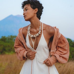 Puede incluir: Una mujer con un vestido blanco con un escote en V profundo y una chaqueta marrón con ribete de volantes. Lleva un collar de cuentas blancas y tiene el pelo corto y oscuro.