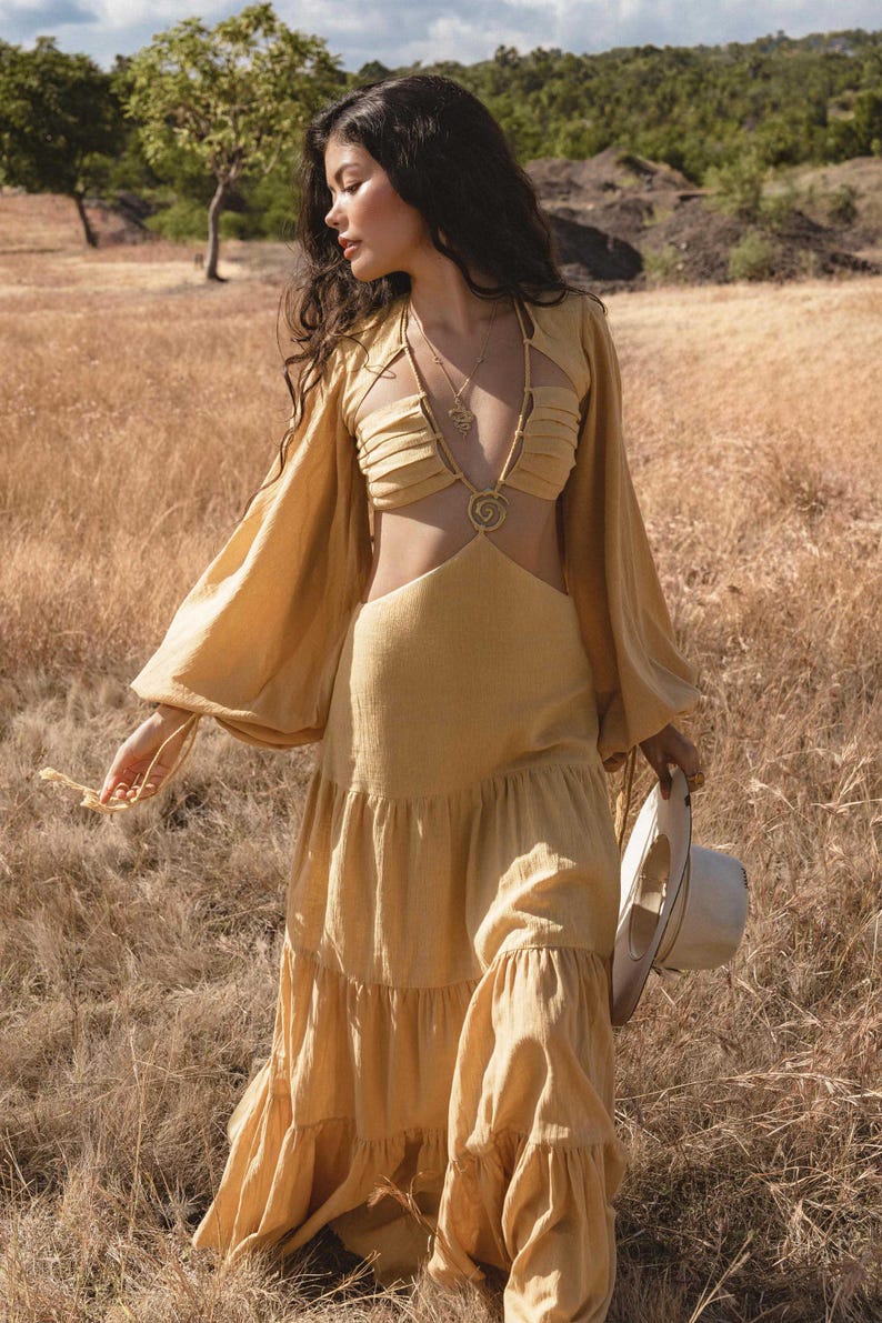 A woman in a flowing Samudra Dress in Saffron Gold by AYA Sacred Wear stands in a dry grassy field, holding a hat, with trees and rocks behind her under a partly cloudy sky. The cotton maxi dress features a tiered bohemian silhouette and waist cut-outs.