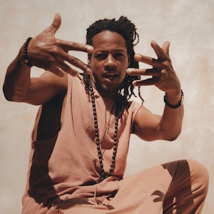 Könnte beinhalten: Ein Mann mit Dreadlocks trägt ein hellbraunes ärmelloses Hemd und eine passende Hose. Er trägt eine Perlenkette und Armbänder an beiden Handgelenken. Er sitzt mit erhobenen Händen vor sich und blickt direkt in die Kamera.