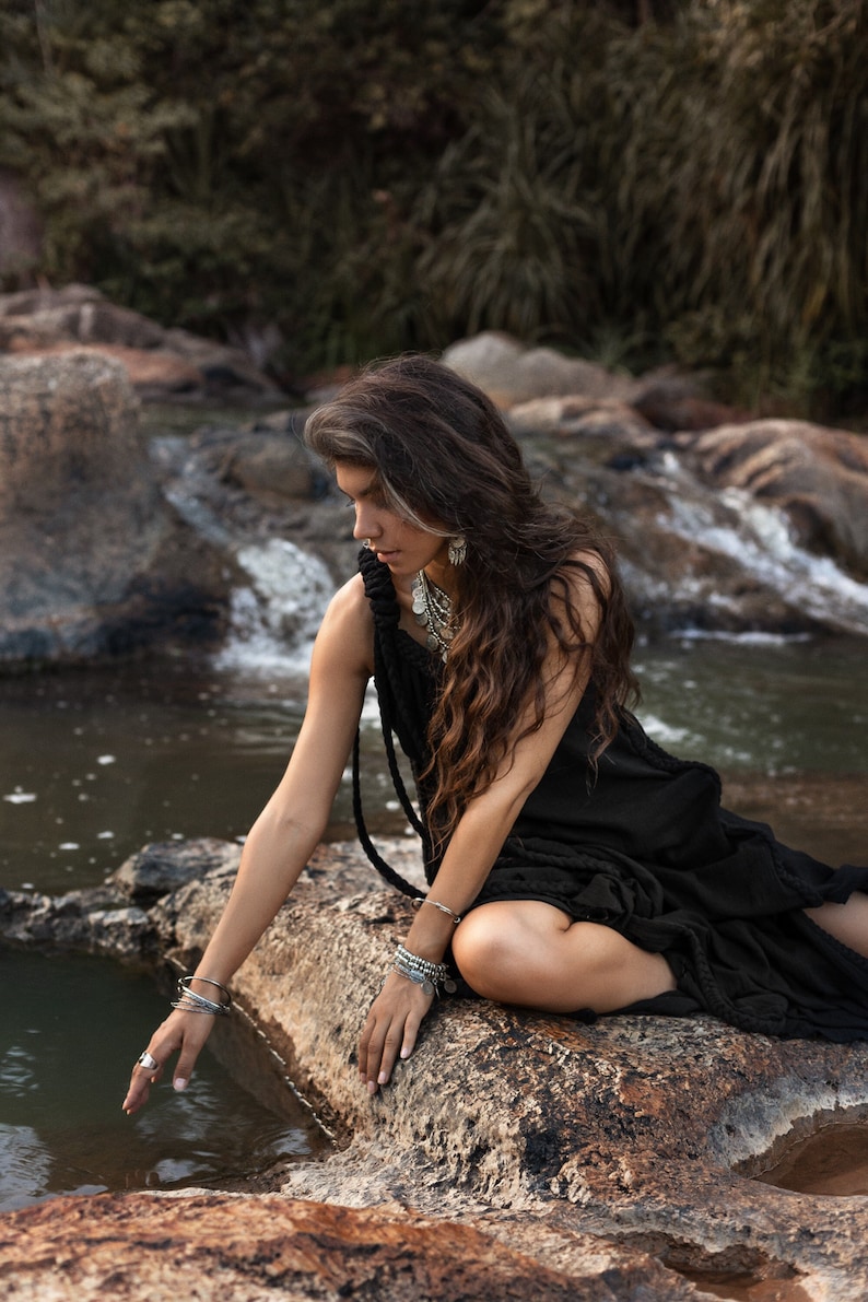 May include: A woman with long brown hair sits on a rock by a stream. She is wearing a black dress and silver jewelry. Her hand is reaching out to the water.