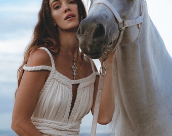 May include: A woman in a white dress with a braided rope design stands with a white horse. She is wearing a silver necklace with a pendant. The horse is wearing a white halter.
