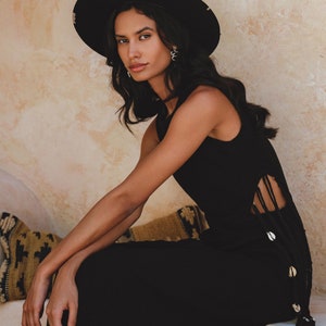 May include: A woman wearing a black sleeveless dress with a side slit and shell accents. She is wearing a black wide-brimmed hat and has long dark hair.
