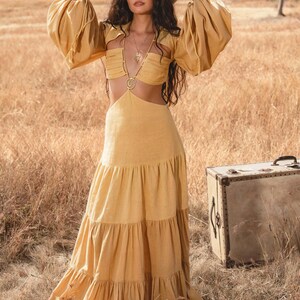A woman in the AYA Sacred Wear Samudra Dress in Saffron Gold, a cotton maxi with a tiered bohemian silhouette and waist cut-outs, stands by a vintage suitcase in a dry field under blue sky, wearing a wide-brimmed hat.