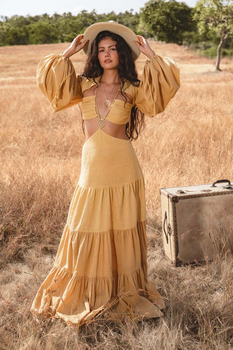 A woman in the AYA Sacred Wear Samudra Dress in Saffron Gold, a cotton maxi with a tiered bohemian silhouette and waist cut-outs, stands by a vintage suitcase in a dry field under blue sky, wearing a wide-brimmed hat.