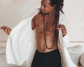 May include: A man with dreadlocks is wearing a white linen shirt and black pants. He is wearing a beaded necklace with a tassel. He is standing in front of a white wall.