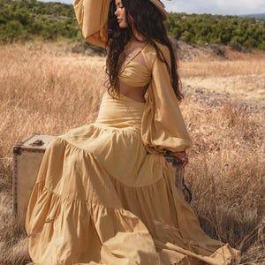 A woman in the Samudra Dress in Saffron Gold by AYA Sacred Wear—an airy cotton maxi with tiered bohemian silhouette and waist cut-outs—sits on a suitcase in a dry grassy field, gazing thoughtfully under a partly cloudy sky.