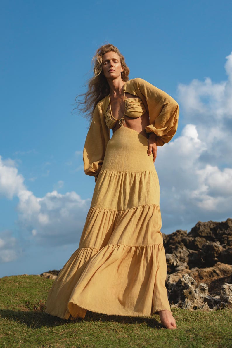 A woman stands barefoot on grass before rocky terrain, wearing the AYA Sacred Wear Samudra Dress in Saffron Gold—a cotton maxi dress with a tiered bohemian silhouette and waist cut-outs—under a blue sky with clouds.