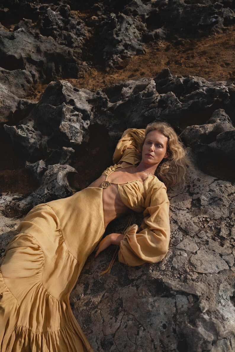 A woman reclines on rocky terrain, gazing at the camera in AYA Sacred Wear’s Samudra Dress in Saffron Gold—a cotton maxi with a tiered bohemian silhouette and waist cut-outs—her wavy hair framing her face against dramatic stone textures.