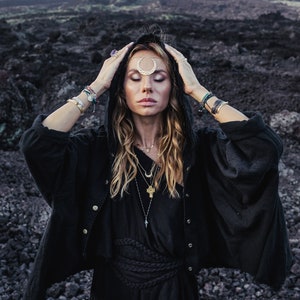 Könnte beinhalten: Eine Frau in einer schwarzen Kapuzenrobe mit einem geflochtenen Gürtel, einem Stirnband in Form eines Halbmondes und mehreren Halsketten. Sie steht in einer felsigen, vulkanischen Landschaft.