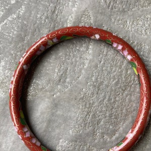 May include: A red cloisonné bangle bracelet with a floral design. The bracelet is decorated with pink and green flowers and gold accents.