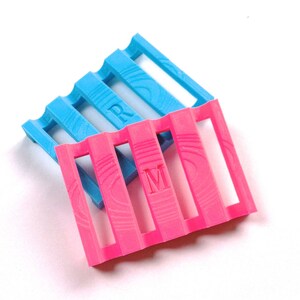 Könnte beinhalten: Zwei farbenfrohe Seifenschalen mit Schlitzen. Eine ist leuchtend rosa mit dem Buchstaben "M" und die andere hellblau mit dem Buchstaben "R". Die Seifenschalen bestehen aus einem strukturierten Material und sind zueinander geneigt.