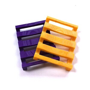 Könnte beinhalten: Zwei kleine, dekorative Gegenstände in Palettenform. Einer ist ein leuchtendes Lila, der andere ein leuchtendes Gelb-Orange. Beide haben ein Latten-Design, das an Miniatur-Holzpaletten erinnert. Die Artikel dienen wahrscheinlich dekorativen Zwecken oder als kleine Tabletts.