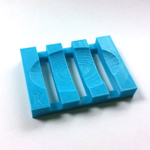 May include: A bright turquoise soap dish with a slotted design. The dish has a rectangular shape with vertical bars and a curved design on the side. The letter "K" is visible on the side of the dish.