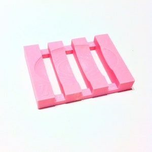 Könnte beinhalten: Eine rosa Seifenschale mit einem geschlitzten Design. Die Schale hat vier erhöhte Stäbe und ein geschwungenes Design an der Seite. Die Seifenschale besteht aus einem festen Material und ist zur Aufnahme eines Seifenstücks bestimmt.