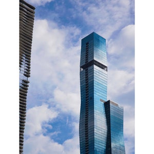 May include: Two tall, modern skyscrapers against a bright blue sky with white clouds. The building on the right is a blue glass tower with a curved design. The building on the left is a gray tower with a wavy design.